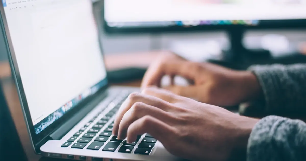 Pessoa digitando em notebook com monitor externo ao fundo em ambiente de home office