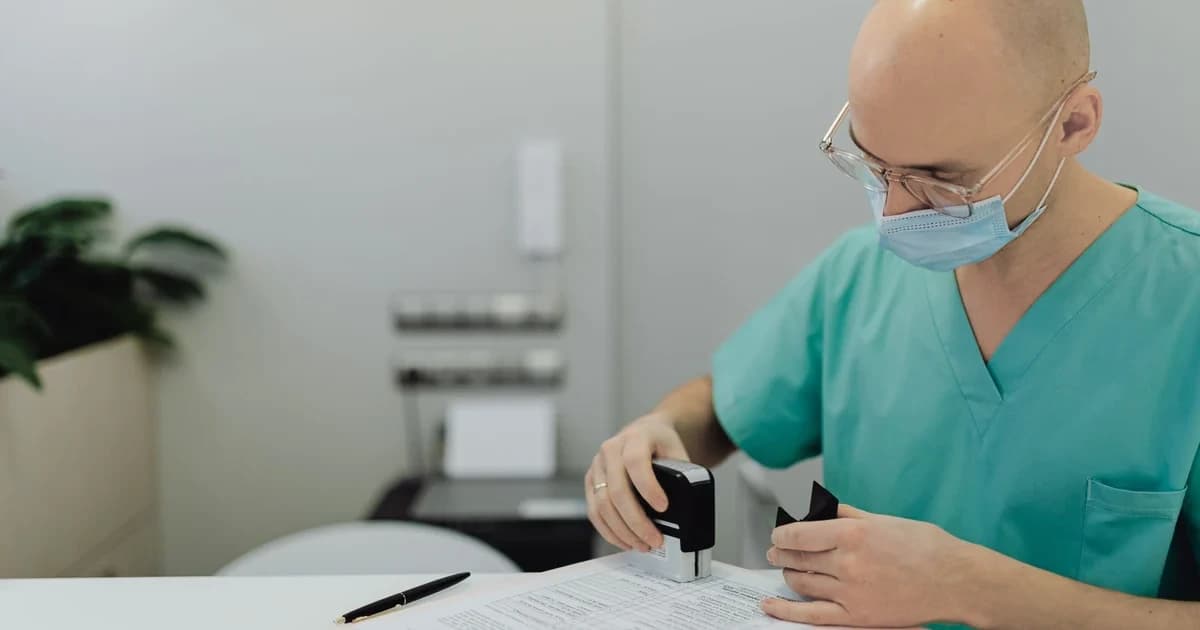 Profissional de saúde de uniforme verde-água aplicando carimbo em um formulário sobre a mesa de um consultório.