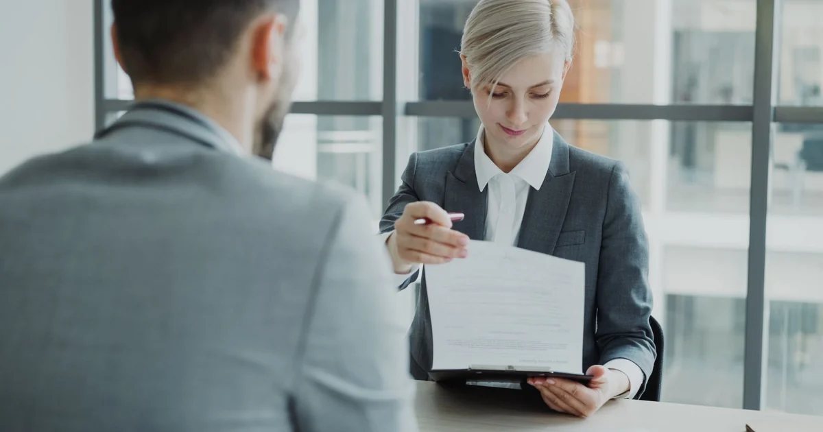 Consultora apresenta um contrato a um cliente do outro lado da mesa, em escritorio claro.