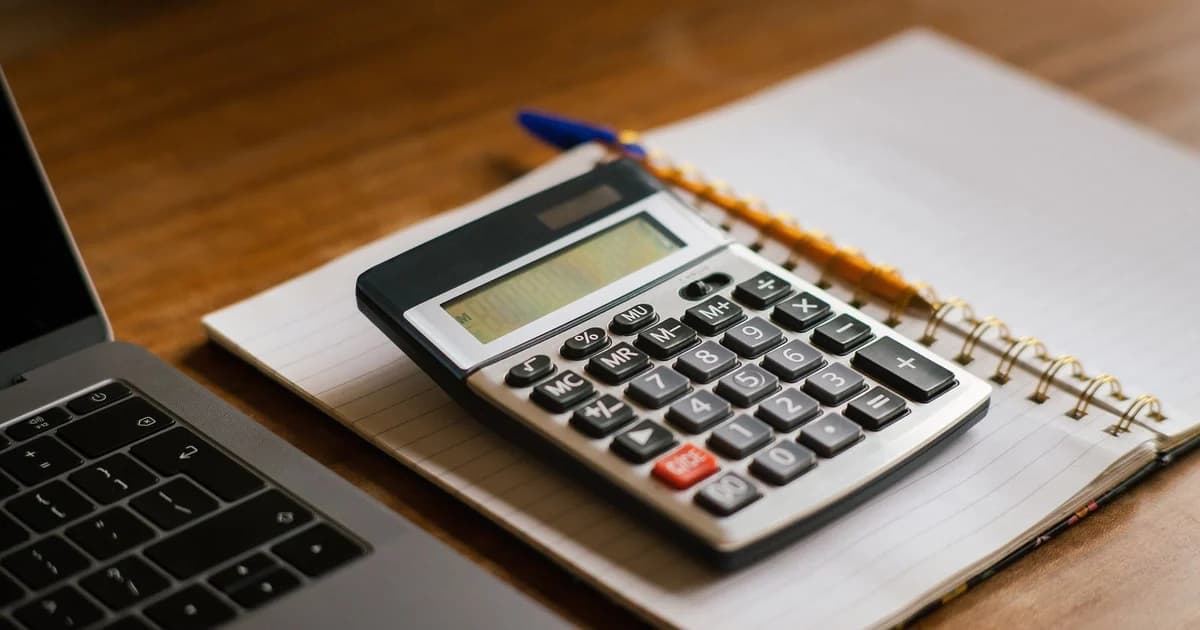 Calculadora de mesa apoiada sobre um caderno pautado aberto, com caneta e lápis ao lado e a borda de um laptop à esquerda, sobre mesa de madeira; nenhuma pessoa e nenhuma cédula no quadro