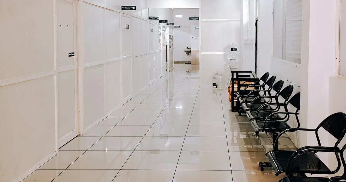 Corredor vazio de uma clínica, com piso claro refletindo a luz e uma fileira de cadeiras metálicas escuras desocupadas junto à parede