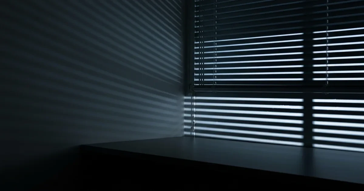 Persiana horizontal entreaberta à noite projetando listras de luz fria azulada sobre parede cinza e mesa escura vazia, sem pessoas