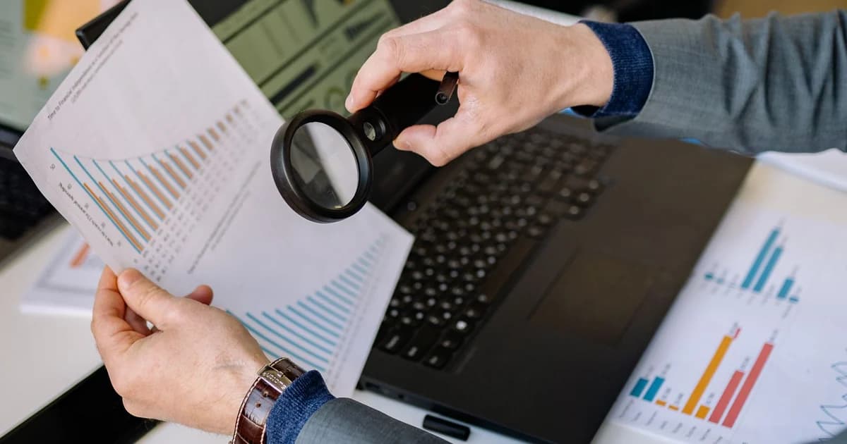 Mãos examinando com lupa um relatório impresso de gráficos diante de um notebook
