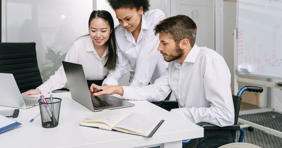 Equipe diversa reunida em torno de uma mesa de escritório com notebook, incluindo colega em cadeira de rodas