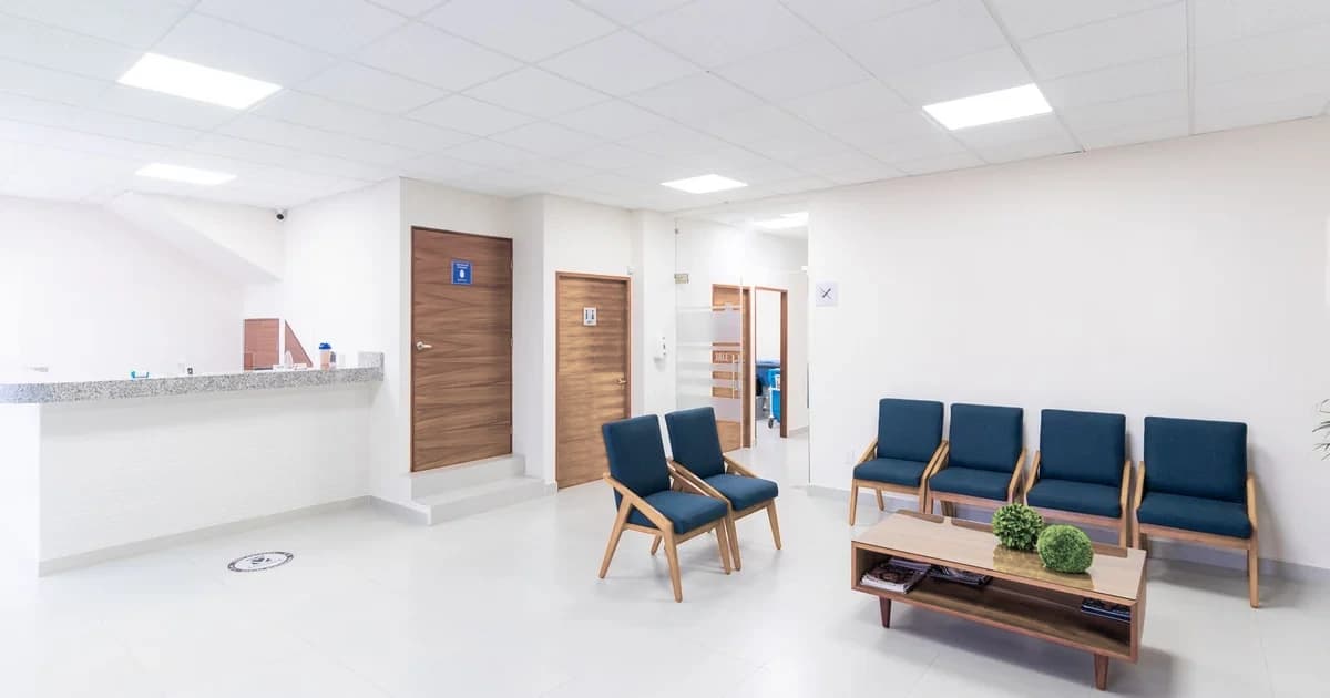 Sala de espera vazia de um hospital, com balcão de recepção à esquerda e cadeiras azuis alinhadas junto à parede.